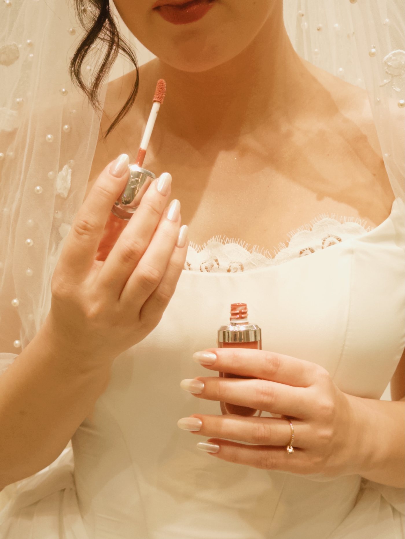 Detalhe da produção de uma noiva: gloss e perfume nas mãos, com o vestido ao fundo